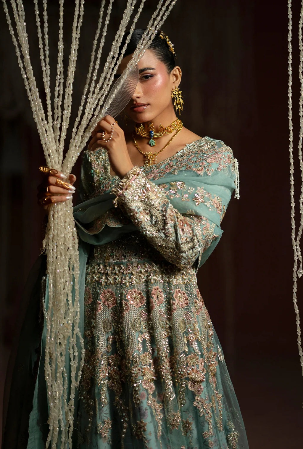 A woman models a traditional Indian outfit consisting of a dusty teal green anarkali kurta and dupatta, heavily embellished with intricate gold and silver embroidery. She is accessorized with gold jewelry and holds a sheer curtain decorated with hanging beads.