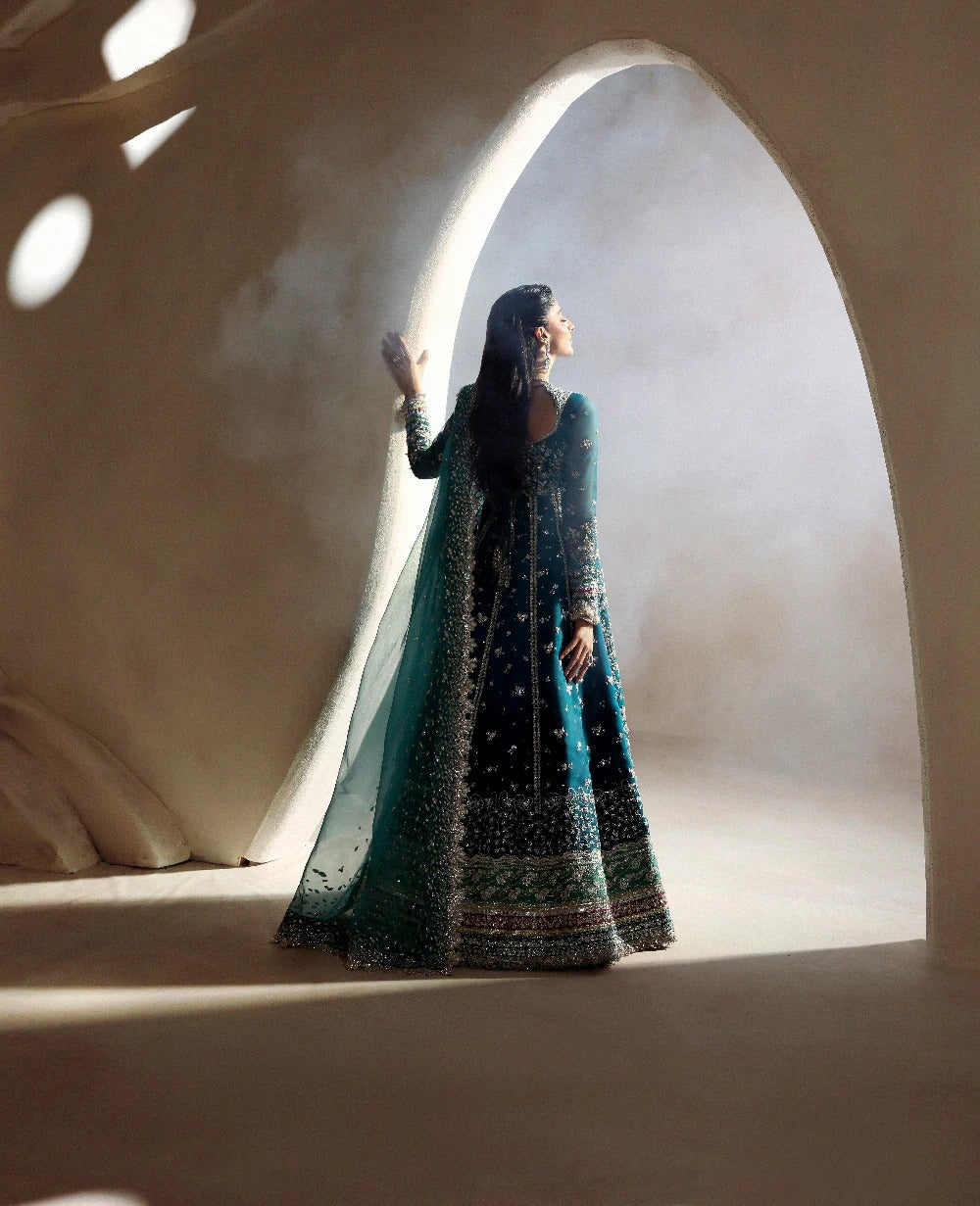 Woman in a long, dark blue embroidered dress standing in a sunlit archway.