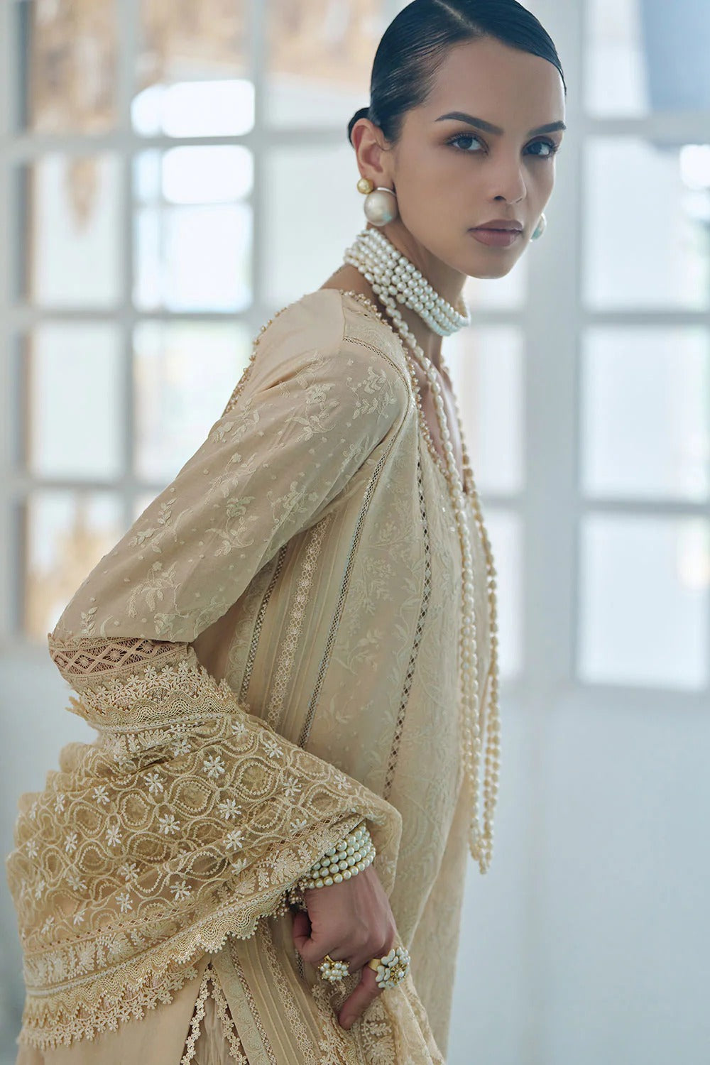 Woman wearing a beige embroidered outfit with pearl accessories in a bright room.