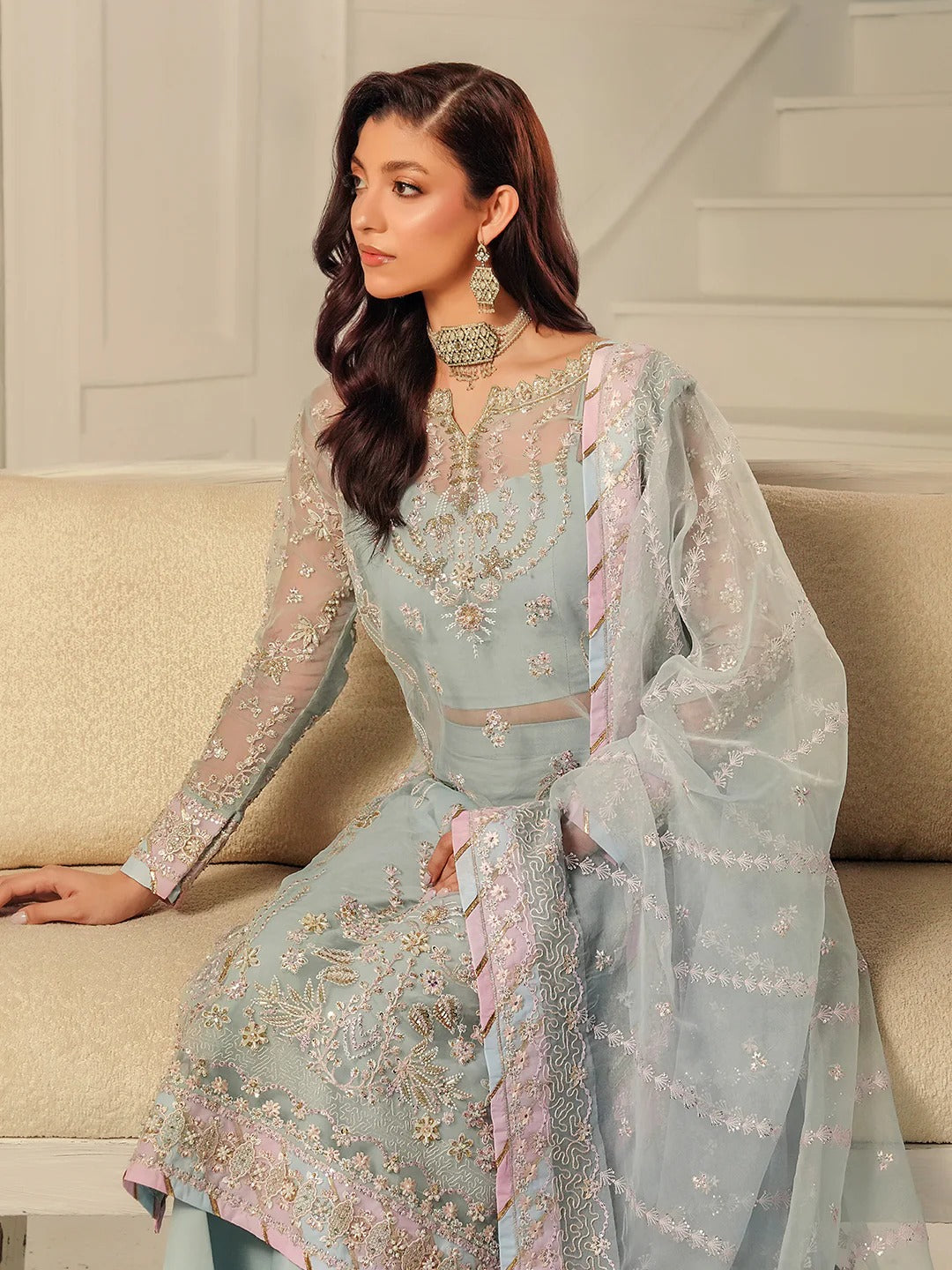Woman in a light blue embroidered traditional outfit sitting on a beige couch.