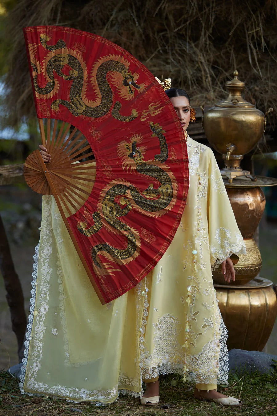 Person in traditional attire holding a red fan with dragon design, standing in a natural setting.