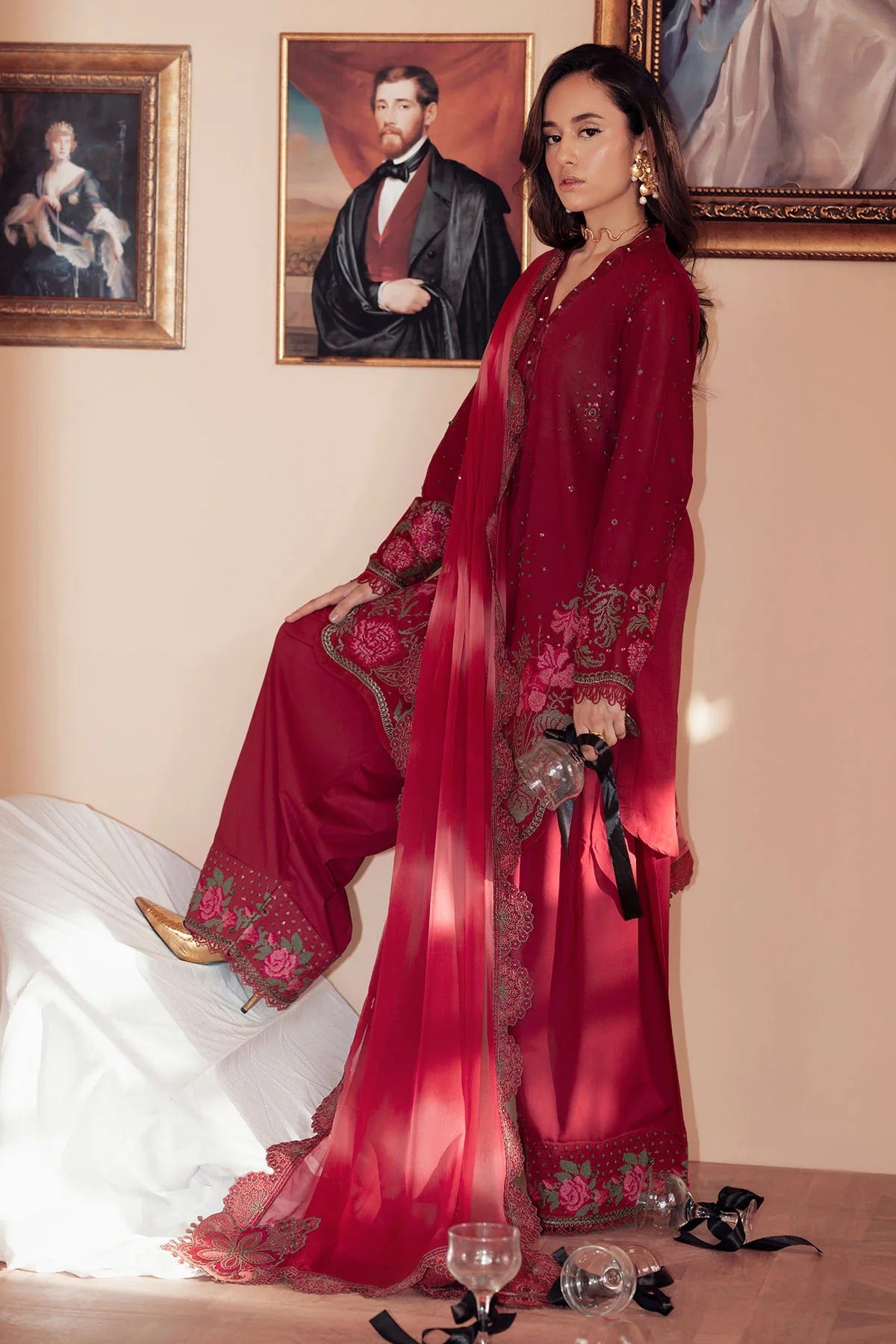 Woman in a red embroidered outfit standing in a room with framed portraits on the wall.