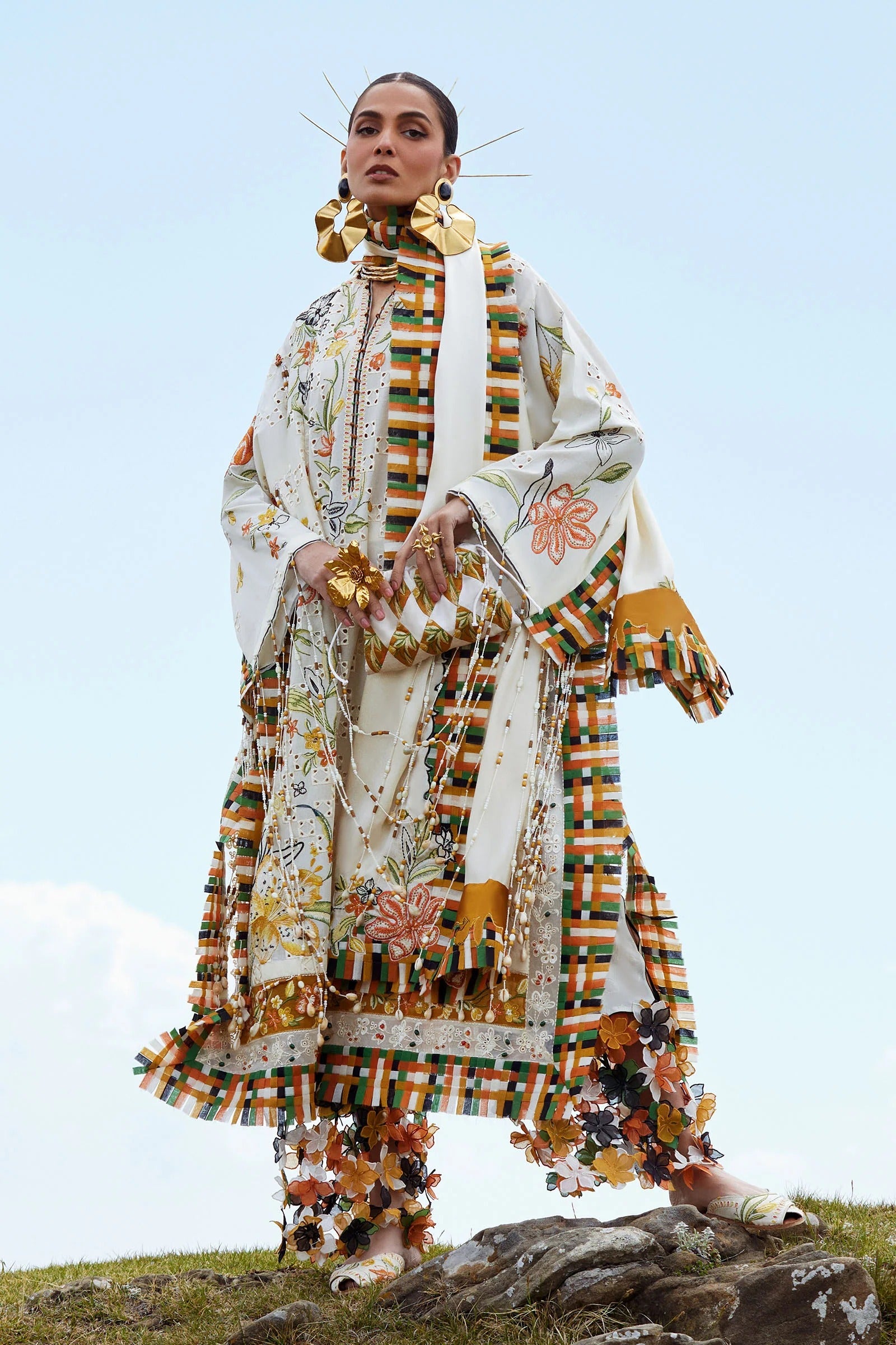 Woman in a colorful, patterned outfit standing on a grassy hill with a clear blue sky.