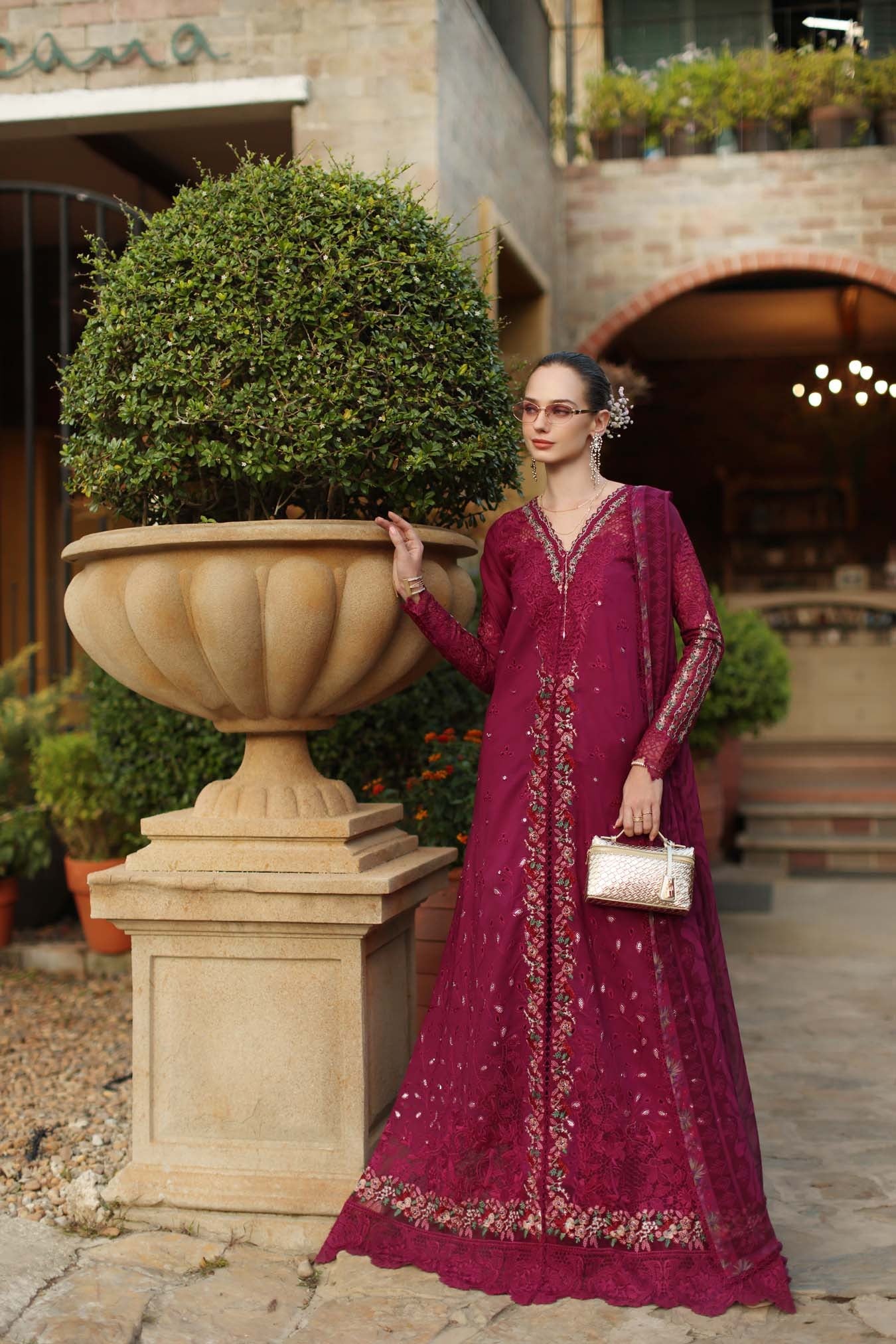 Woman in a burgundy embroidered dress standing next to a large decorative planter outdoors.