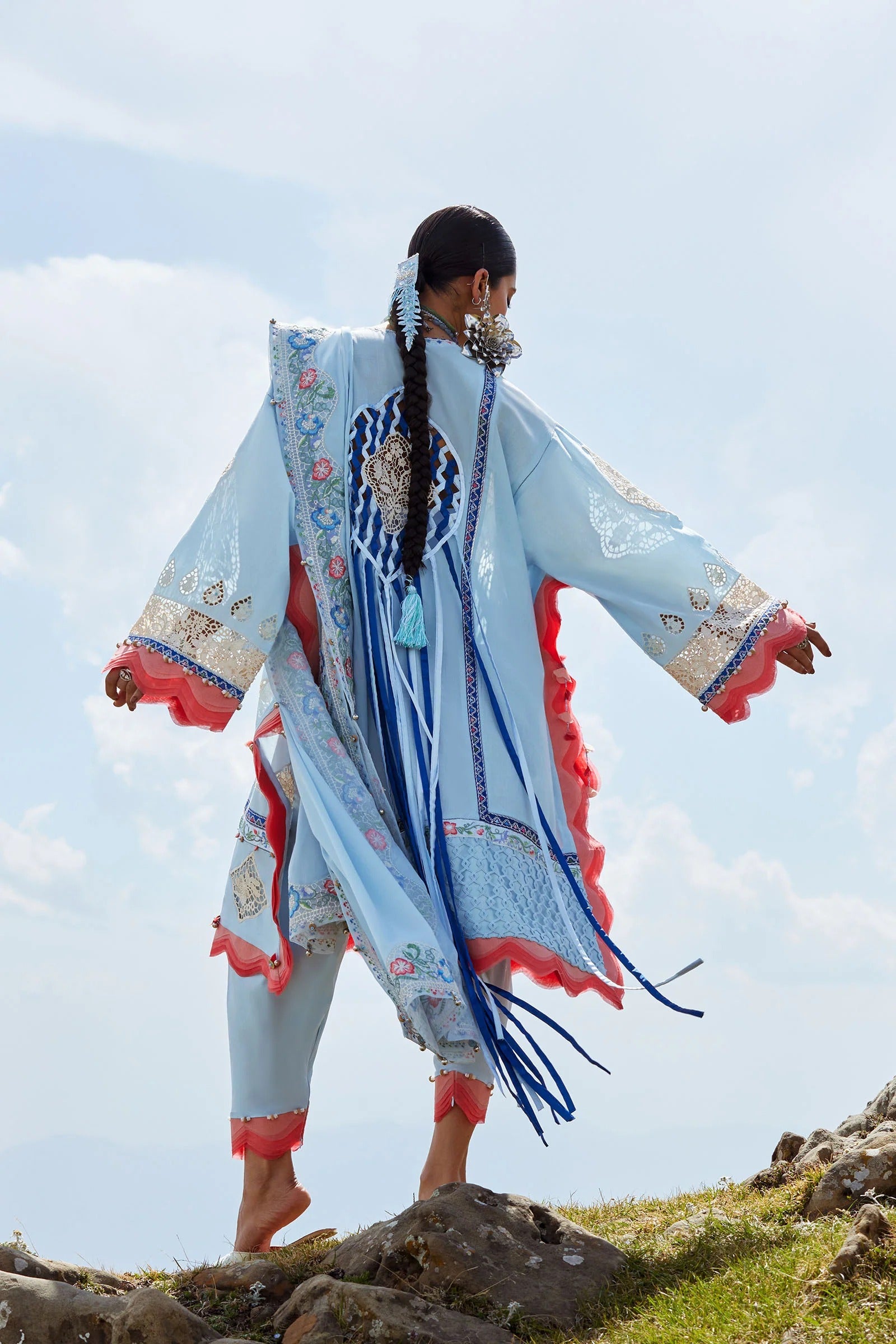 Person wearing a traditional embroidered outfit standing on a rocky landscape with a clear sky.