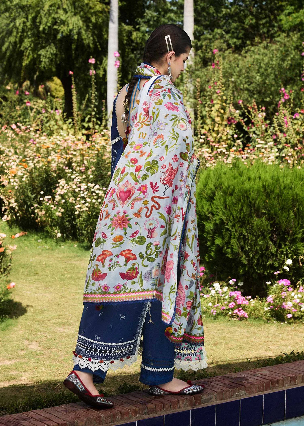 A woman wearing a traditional three-piece salwar kameez suit in a lush garden. The outfit features a white kameez jacket with a vibrant multicolor floral print, navy blue pants with embroidered borders, and a matching navy blue dupatta.