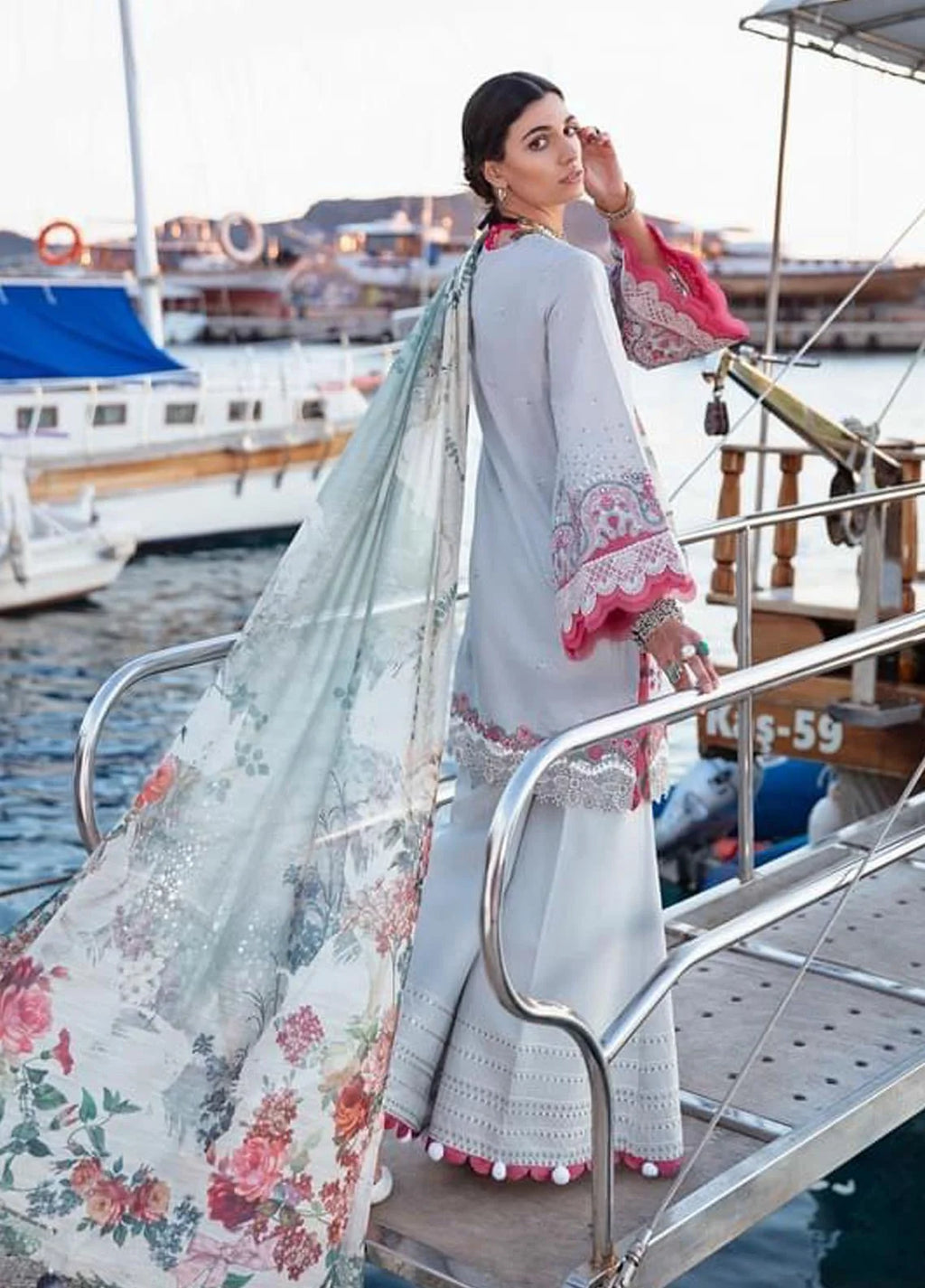 A woman wearing a traditional South Asian outfit stands on a boat deck with water and boats in the background. The outfit consists of a light gray kurta with pink and white embroidered cuffs, a matching light gray dupatta with a pink and green floral print, and a light gray flared skirt with embroidered details and pom-pom trim.