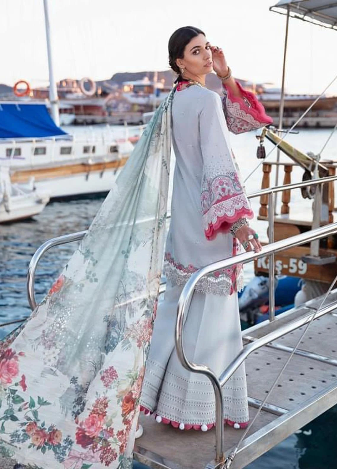 A woman wearing a traditional South Asian outfit stands on a boat deck with water and boats in the background. The outfit consists of a light gray kurta with pink and white embroidered cuffs, a matching light gray dupatta with a pink and green floral print, and a light gray flared skirt with embroidered details and pom-pom trim.