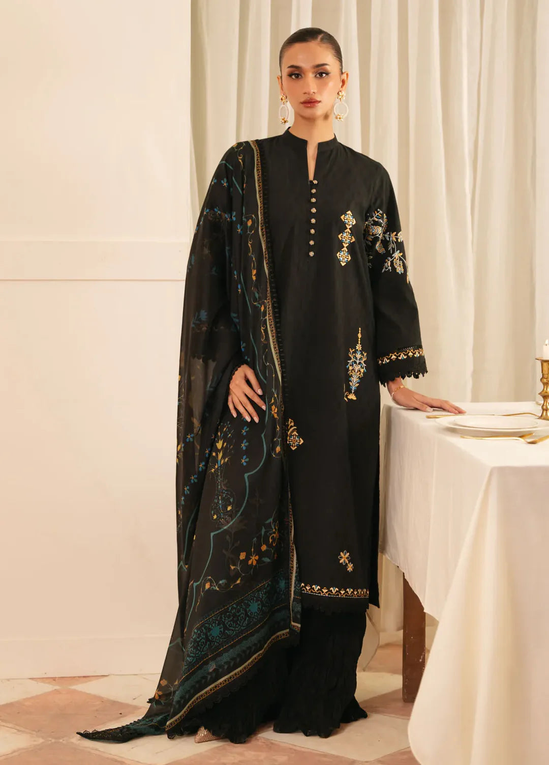 A woman models a black traditional outfit with gold and teal embroidery, featuring a long kurta, matching pants, and a sheer embroidered dupatta, set against a warm cream-colored indoor background.