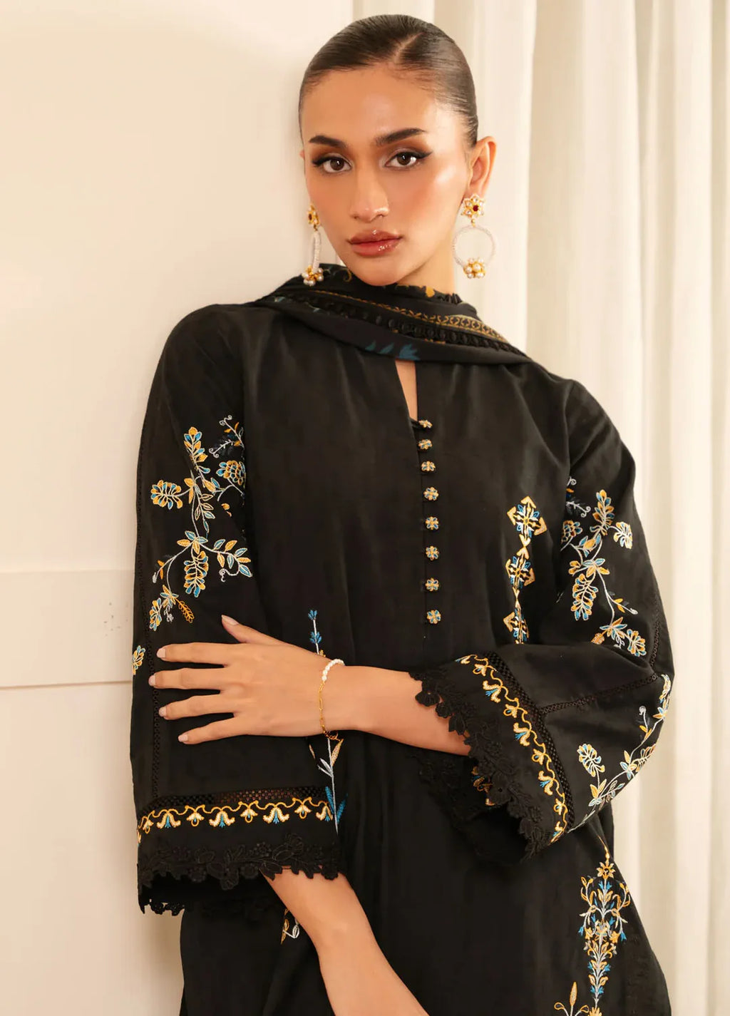 A woman wearing a black embroidered kurta with gold and blue floral detailing, paired with a matching black dupatta, set against a neutral cream-colored background.