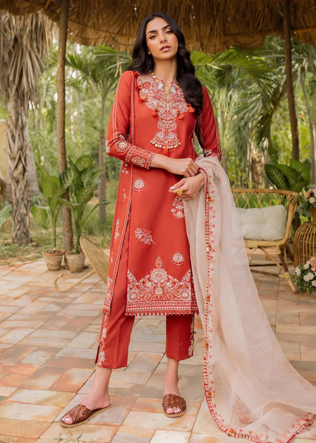 A woman models a traditional three-piece salwar kameez suit in terracotta red with intricate white embroidery, paired with matching cropped trousers and a sheer beige dupatta, set against a tropical garden patio background.