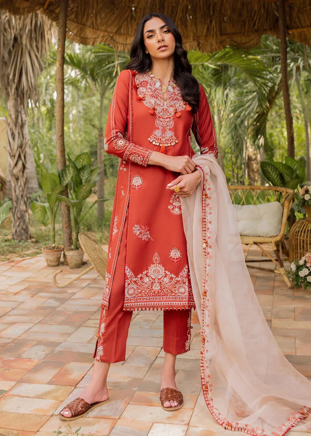 A woman models a traditional three-piece salwar kameez suit in terracotta red with intricate white embroidery, paired with matching cropped trousers and a sheer beige dupatta, set against a tropical garden patio background.