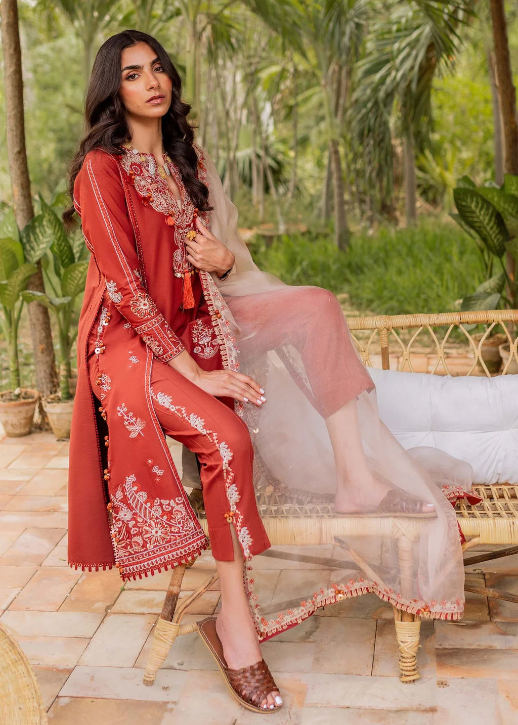 A woman models a rust-red embroidered traditional outfit with matching pants and a sheer peach dupatta, seated on a rattan bench in a tropical garden setting.