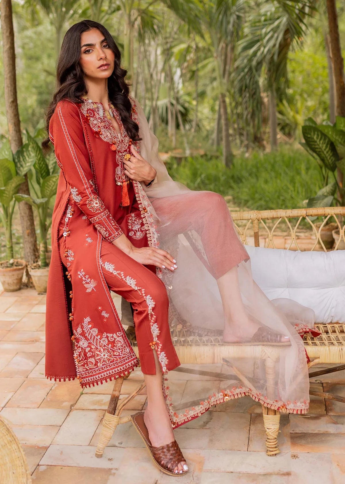 A woman models a rust-red embroidered traditional outfit with matching pants and a sheer peach dupatta, seated on a rattan bench in a tropical garden setting.