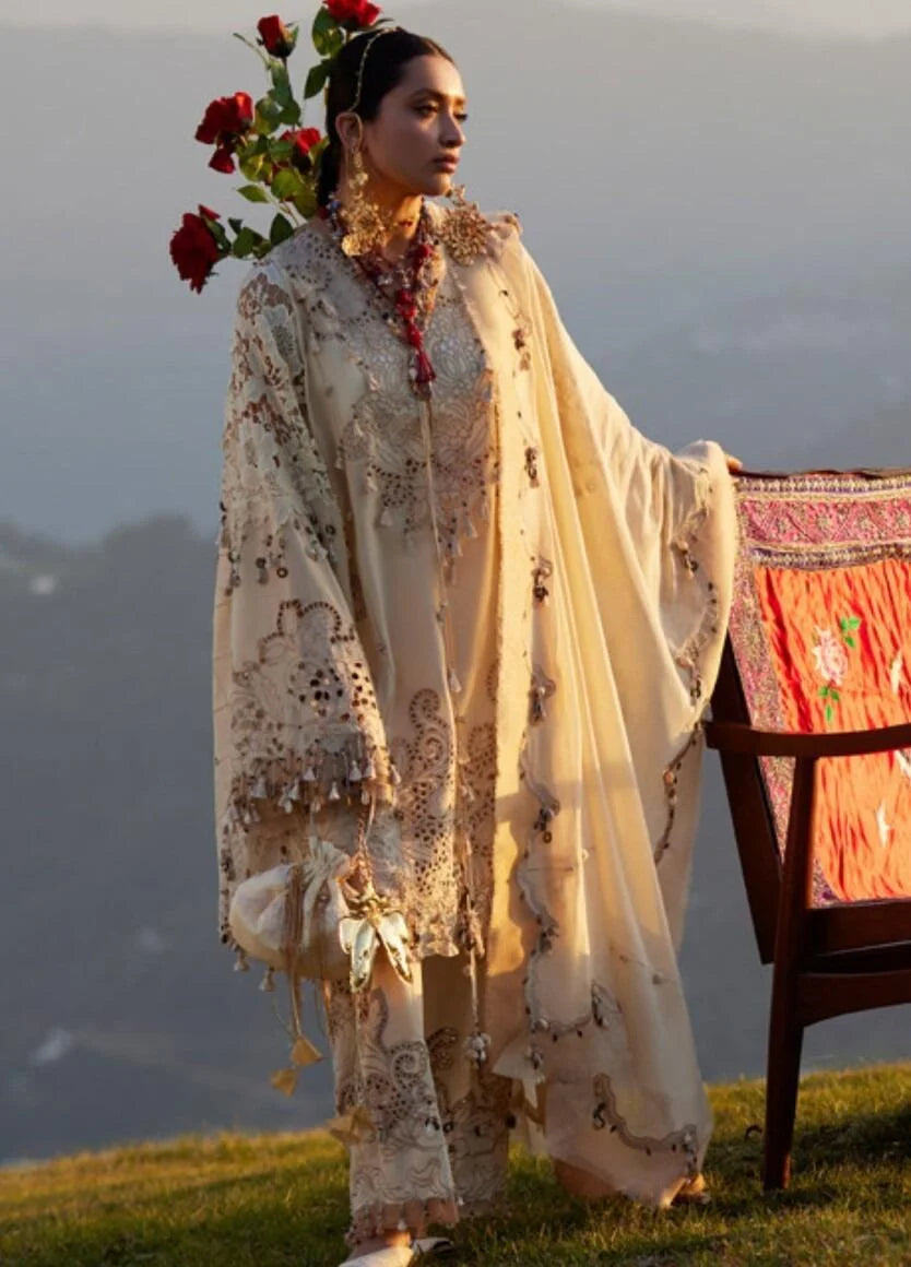 A woman stands outdoors on a grassy hill wearing a flowing cream-colored embroidered traditional outfit with a sheer draped overlay, floral embroidery, and long wide sleeves. Red roses are tucked behind her head, and a wooden chair with a patterned textile sits beside her against a soft-focus mountain background.