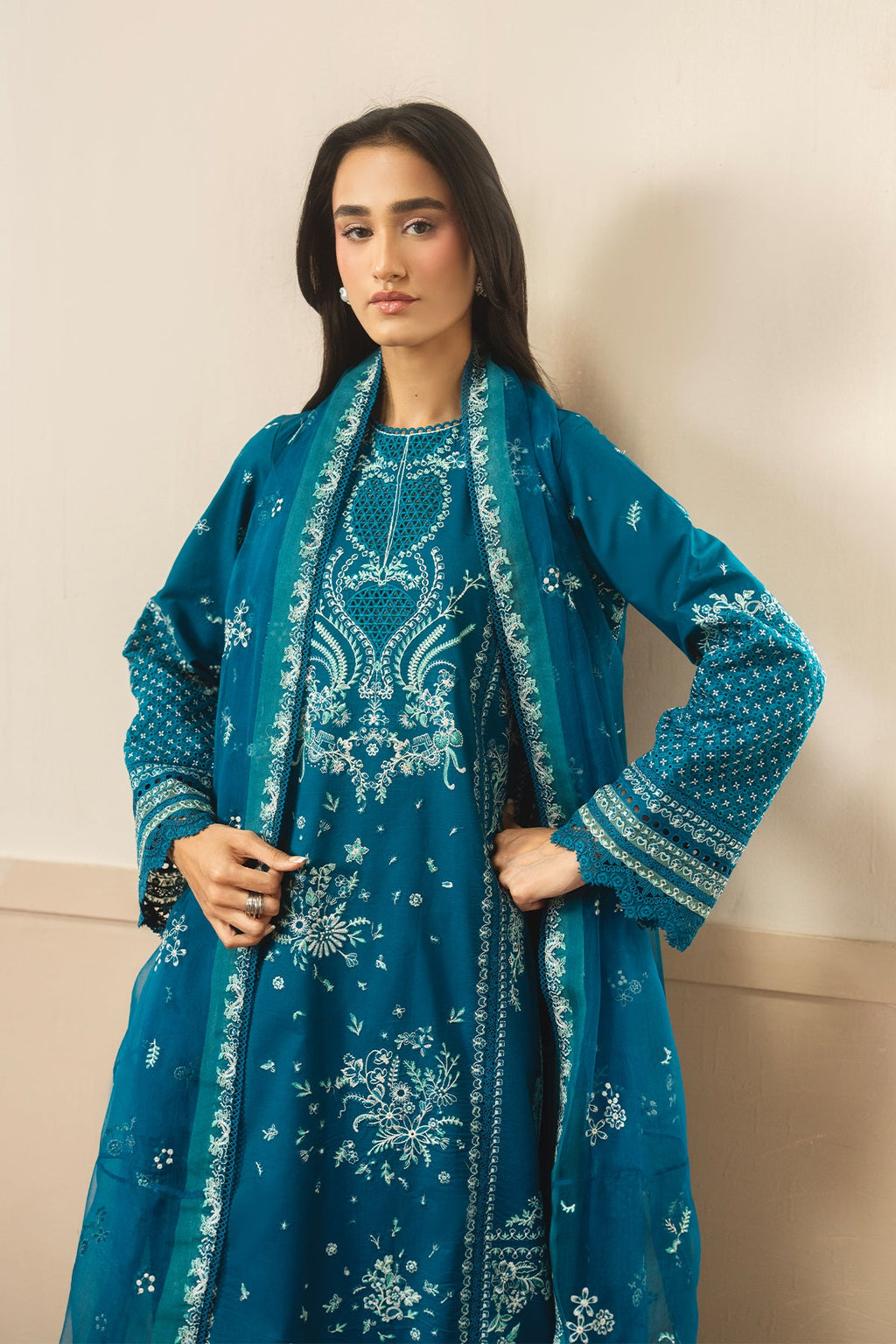 Woman wearing a teal embroidered outfit with a matching shawl against a beige background