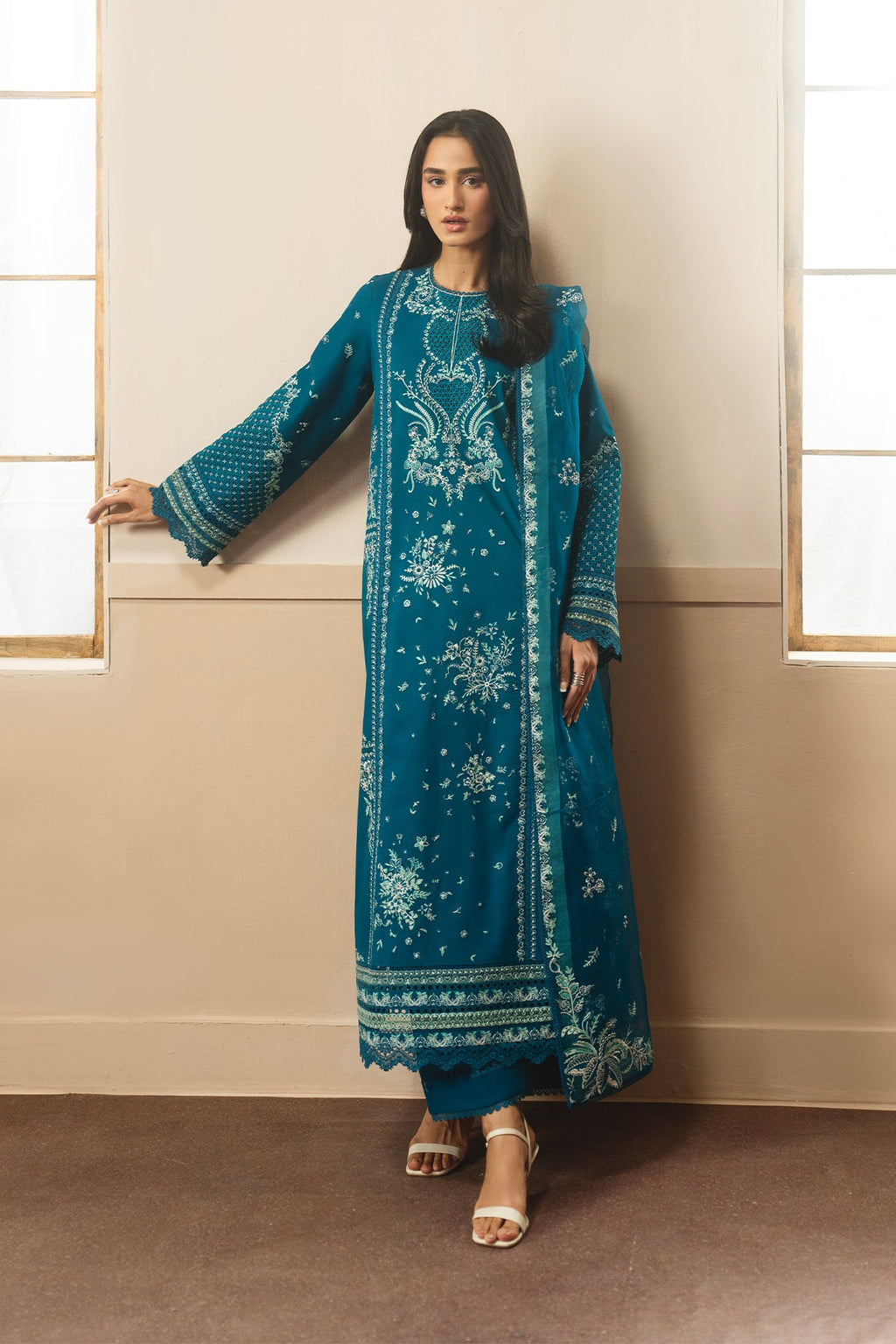 Woman wearing a teal embroidered traditional outfit standing against a beige wall.