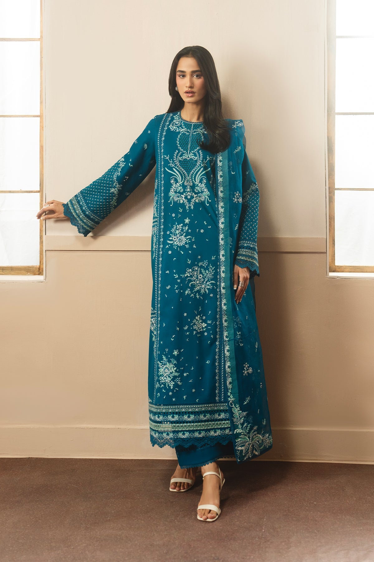 Woman wearing a teal embroidered traditional outfit standing against a beige wall.