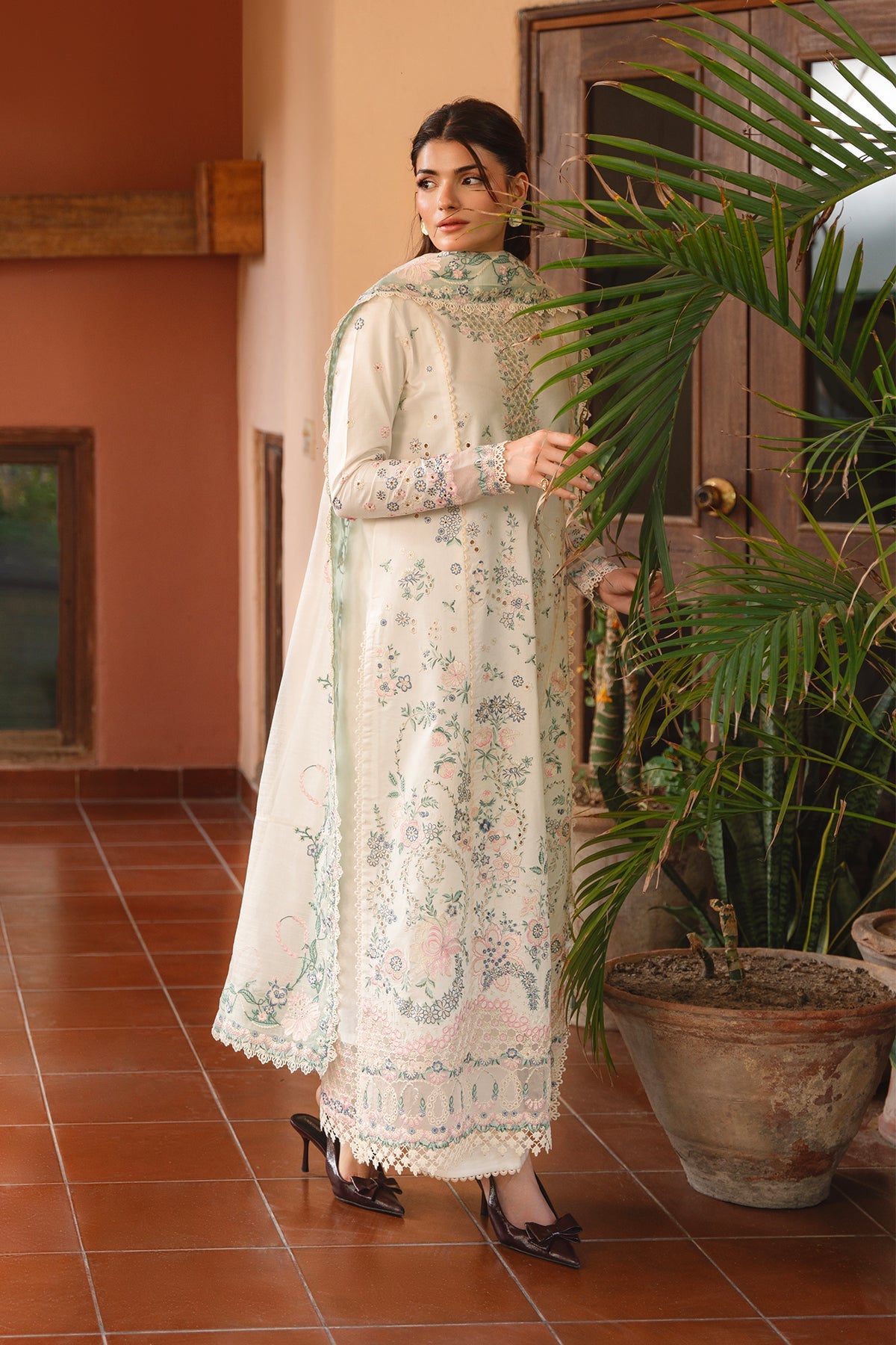 Woman in a floral dress standing in a room with plants and tiled floor.