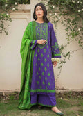 A woman stands outdoors wearing a traditional three-piece salwar kameez suit. The kurta is purple with green floral embroidery, paired with matching purple pants and a bright green dupatta with green patterns.