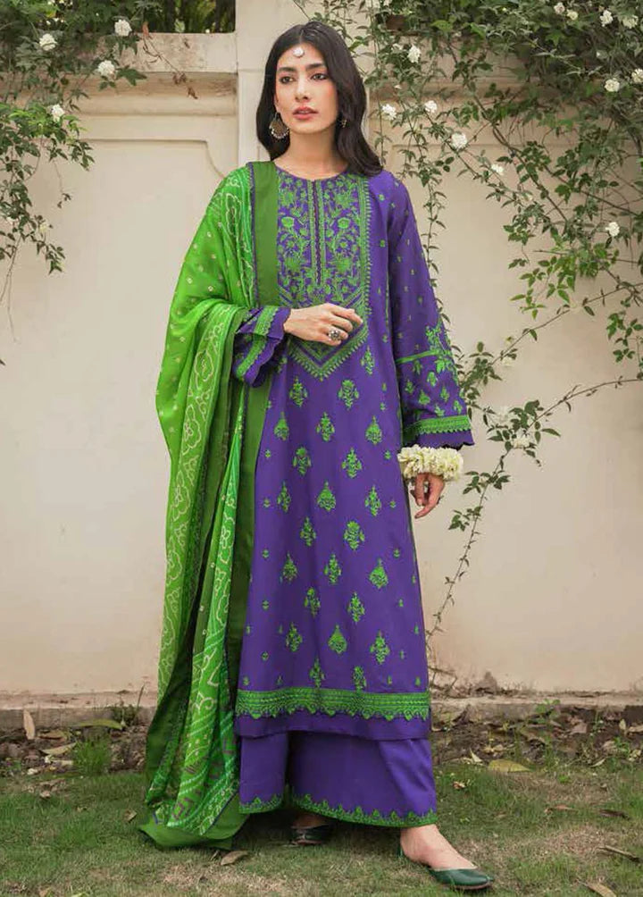 A woman stands outdoors wearing a traditional three-piece salwar kameez suit. The kurta is purple with green floral embroidery, paired with matching purple pants and a bright green dupatta with green patterns.