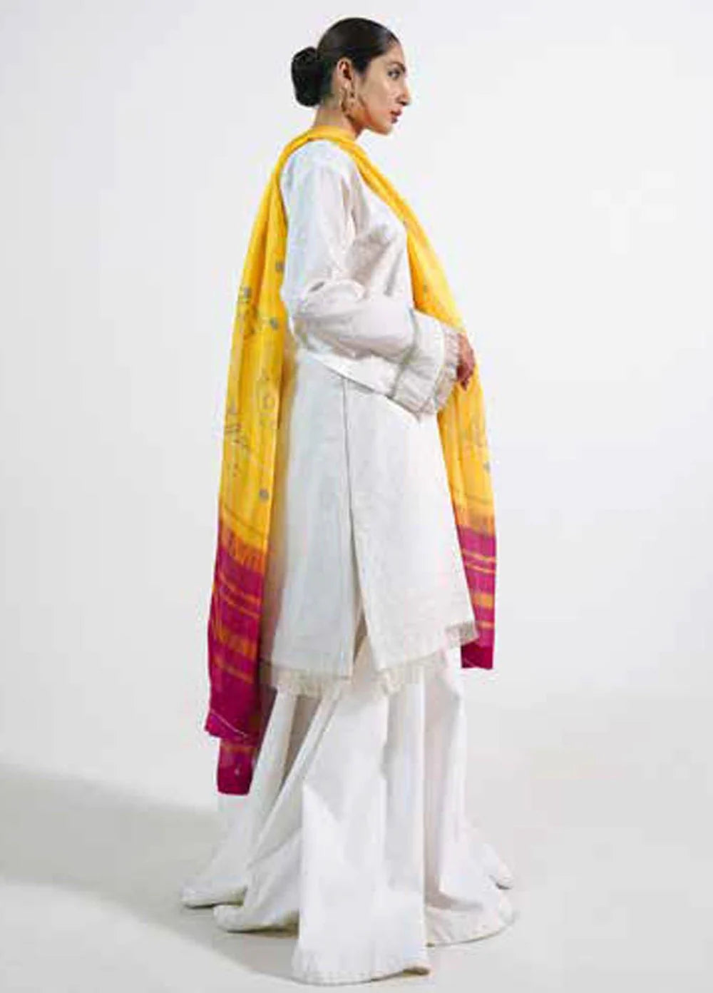 A woman modeling a white traditional outfit with a bright yellow dupatta featuring magenta and yellow striped borders, photographed against a plain light gray studio background.